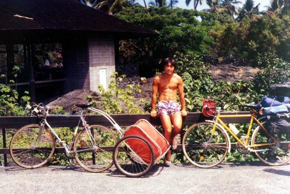 A young man in swim shorts sits on a wooden railing between two loaded touring bicycles and a trailer, tropical trees behind him.