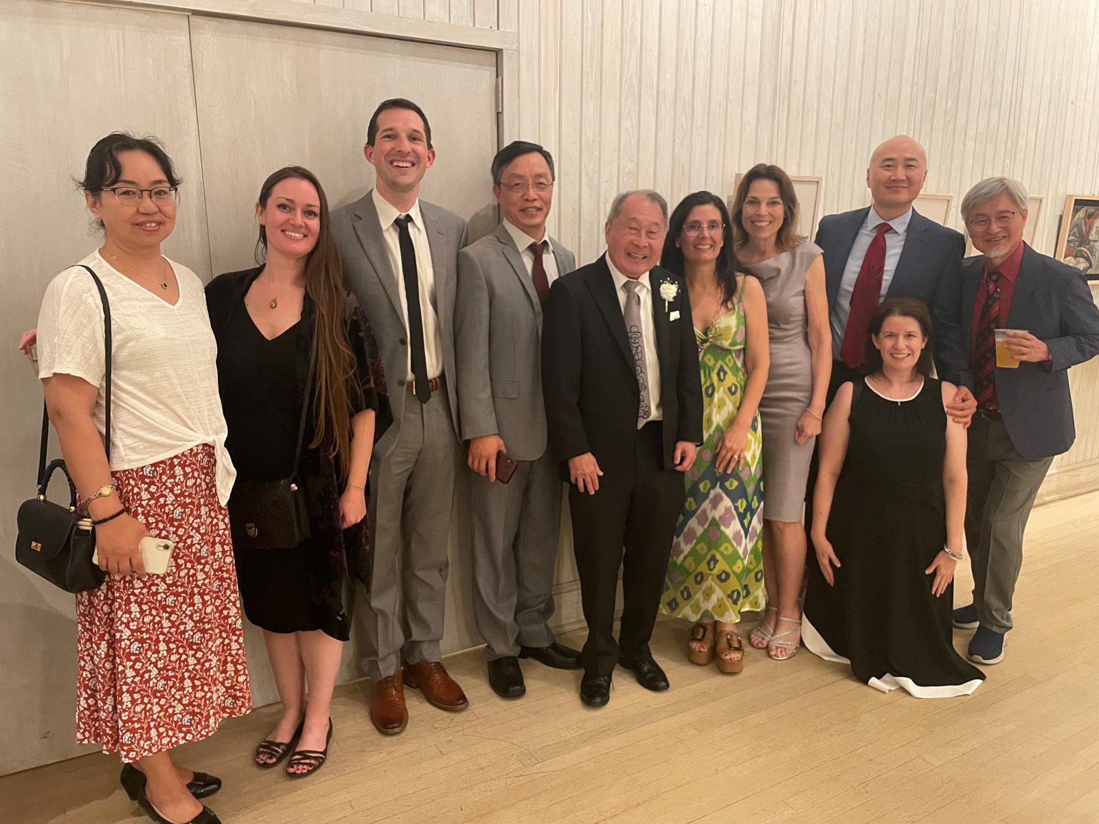 Eleven people in formal clothes pose in a row against a pale wall, Ken Muneoka at the center.