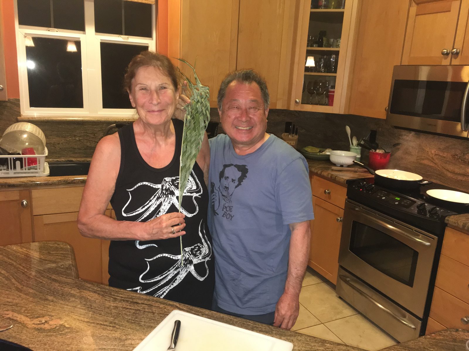Ken Muneoka and Susan Bryant smile side by side in a kitchen; she holds a woven palm frond.
