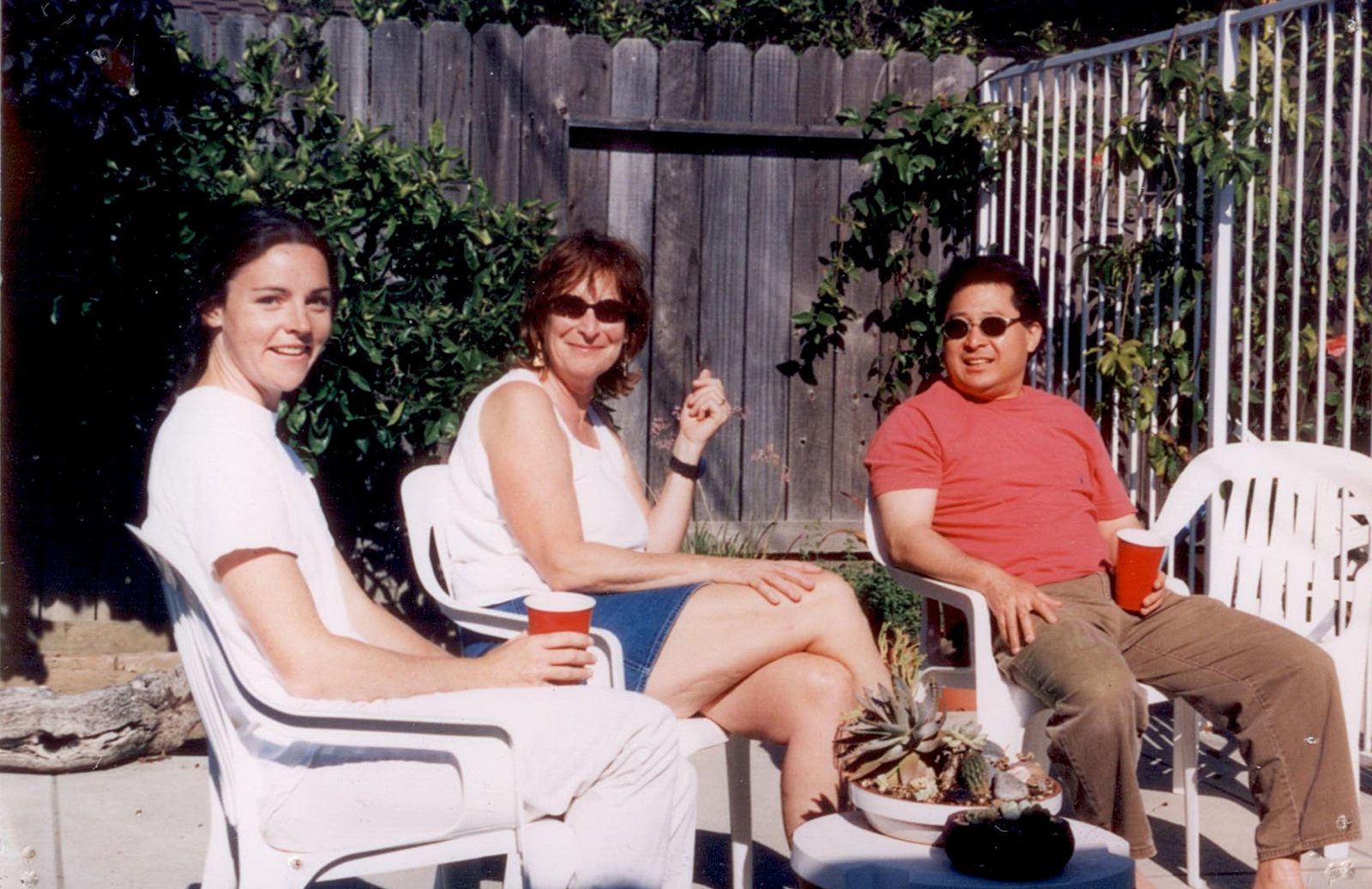 Three people sit in white patio chairs by a wooden fence, two of them holding red cups.